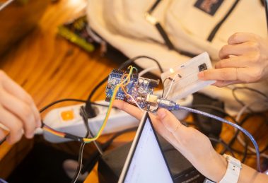 Equipe trabalha em conjunto em um projeto de Arduino, com fios e placa de prototipagem sobre a mesa.