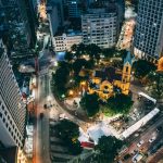 Vista aérea noturna de uma cidade, com uma praça central e uma igreja iluminadas.