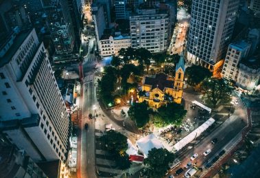 Vista aérea noturna de uma cidade, com uma praça central e uma igreja iluminadas.