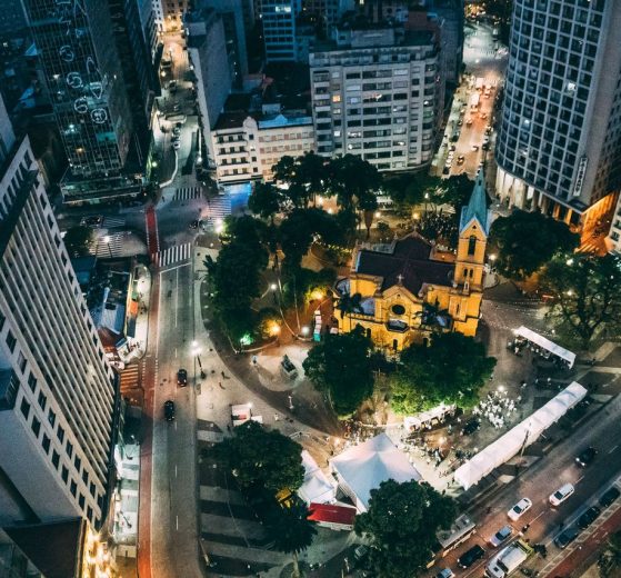 Vista aérea noturna de uma cidade, com uma praça central e uma igreja iluminadas.