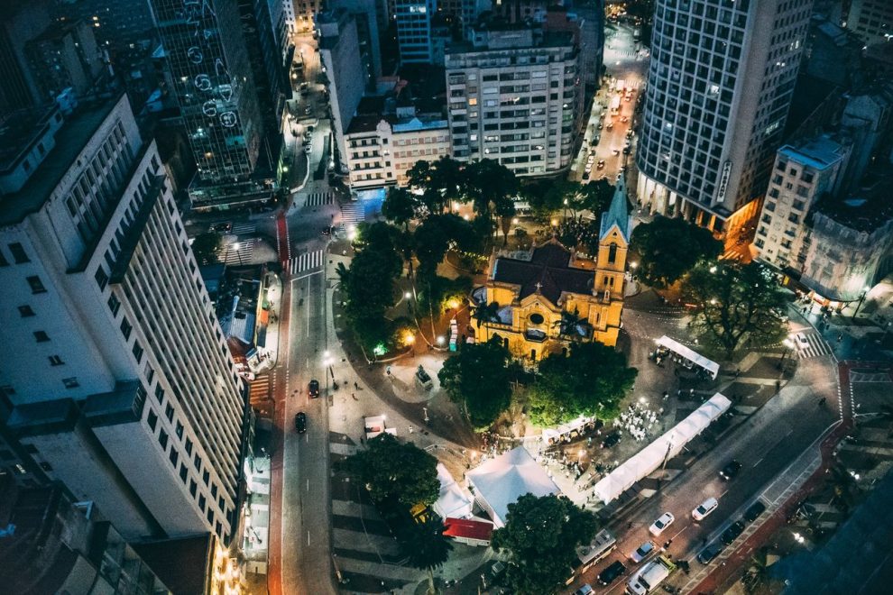 Vista aérea noturna de uma cidade, com uma praça central e uma igreja iluminadas.