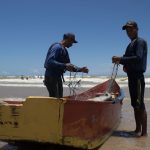 Dois pescadores preparam redes junto a um barco colorido na praia de Itaúnas, no Brasil.