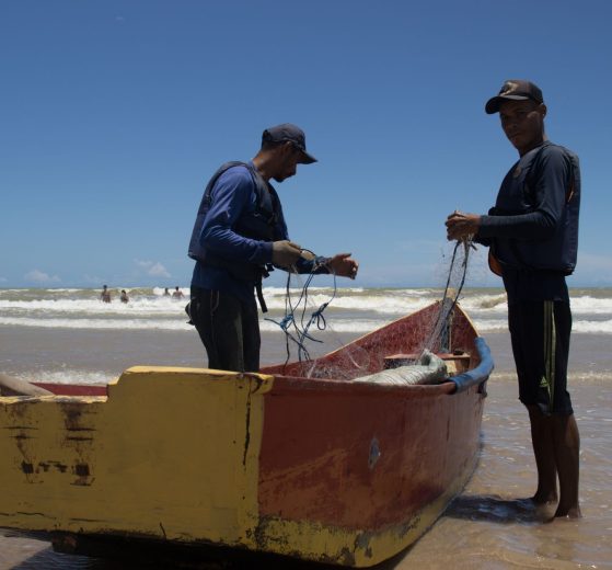 Dois pescadores preparam redes junto a um barco colorido na praia de Itaúnas, no Brasil.