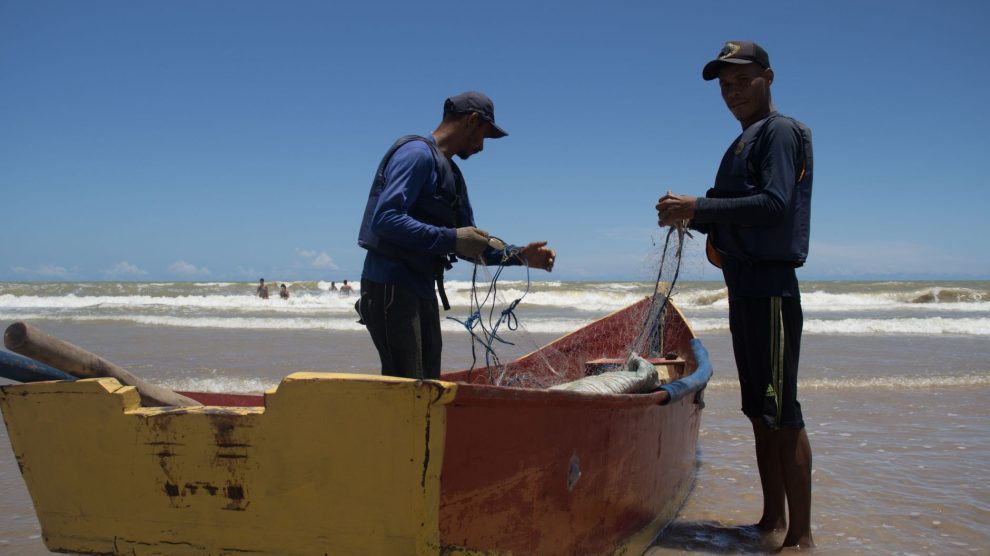 Dois pescadores preparam redes junto a um barco colorido na praia de Itaúnas, no Brasil.