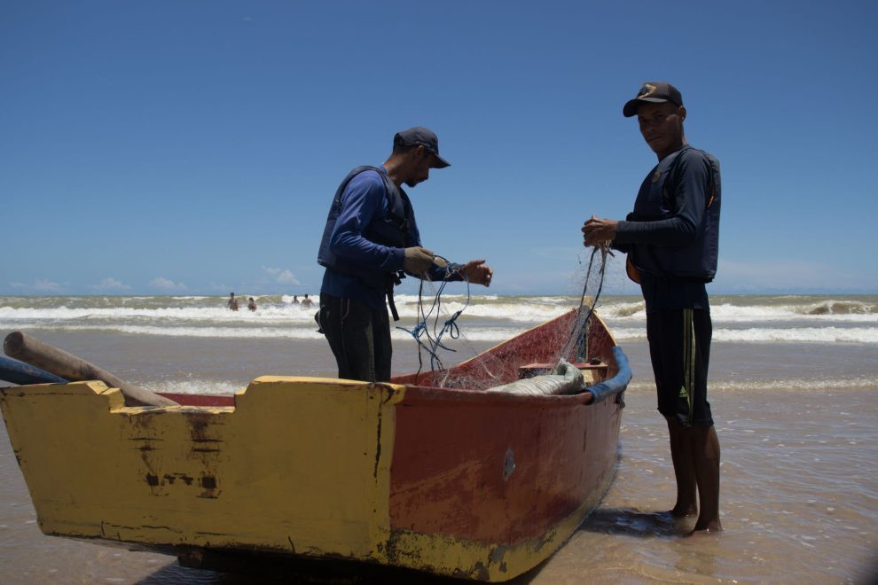 Dois pescadores preparam redes junto a um barco colorido na praia de Itaúnas, no Brasil.